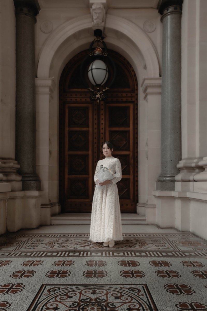 The bride stands small before a towering arched timber door, bouquet in hand