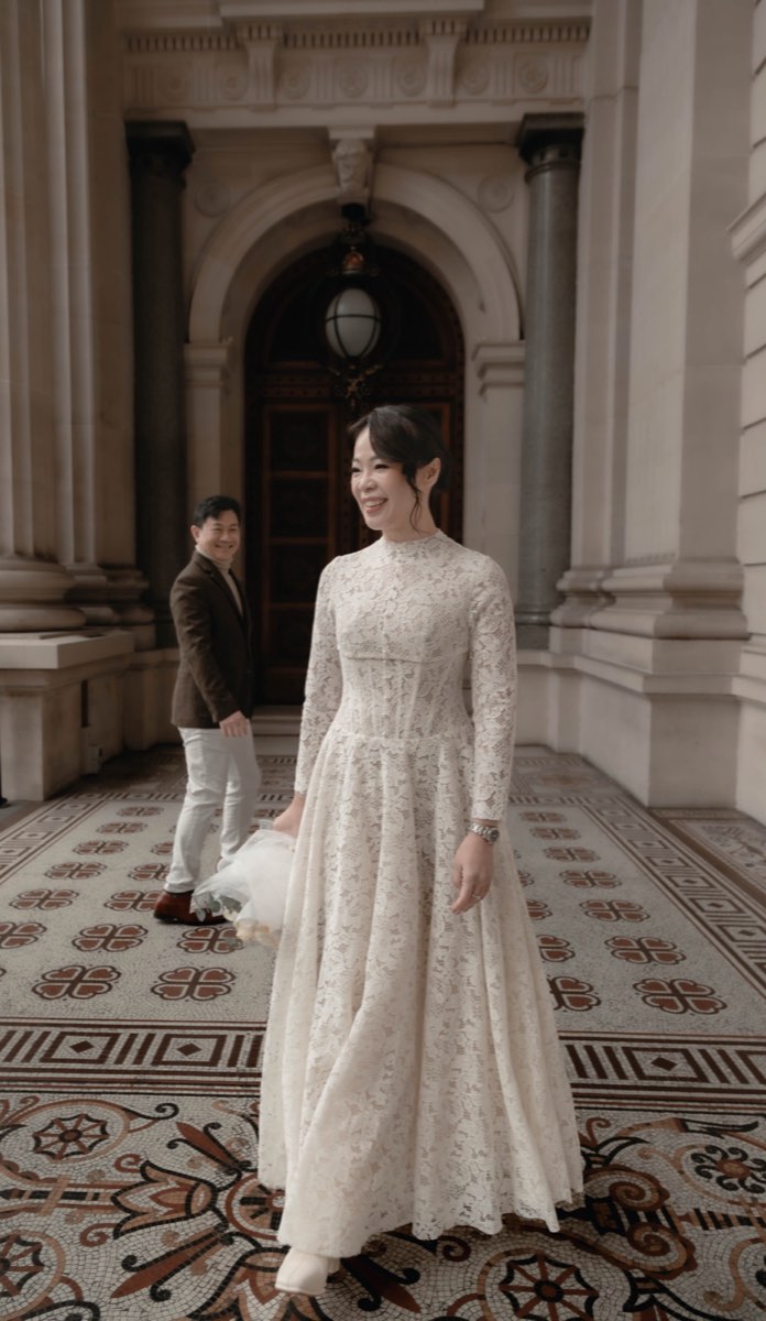 The bride walks the tiled colonnade with her dress fanned out, the groom watching from the columns behind