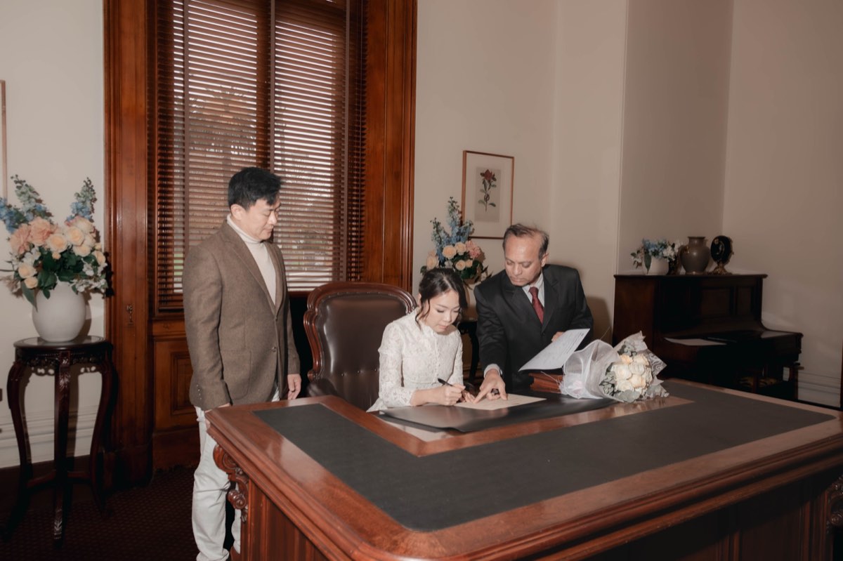 The couple sign the marriage register at a leather topped desk, flowers either side