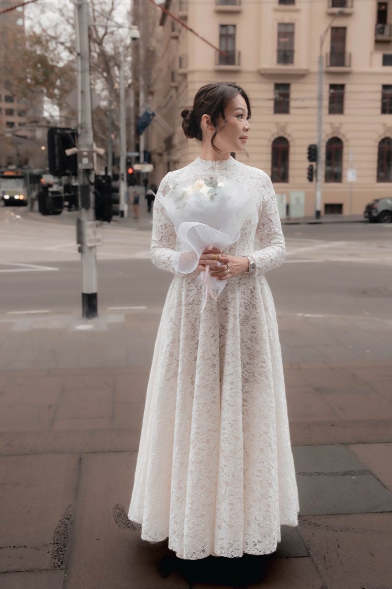The bride stands on a wide Melbourne street holding her bouquet, city behind her
