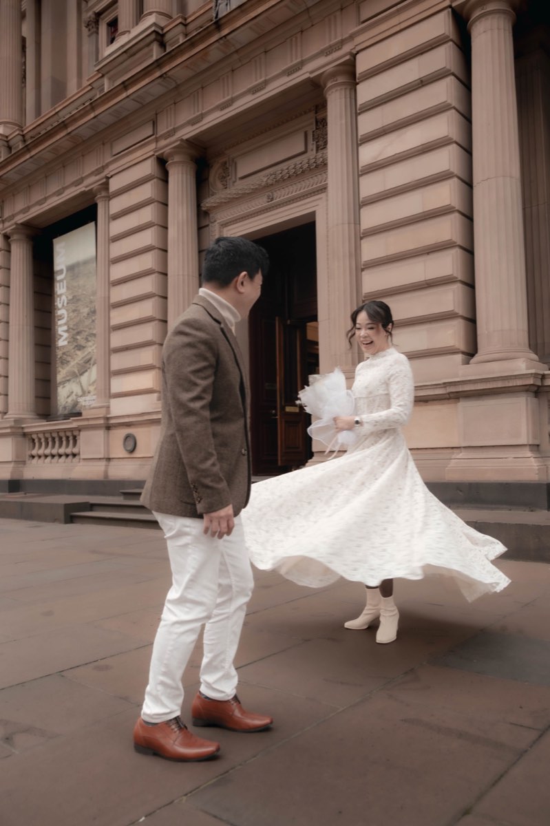 The bride spins her lace dress on the footpath outside the Old Treasury as the groom looks on